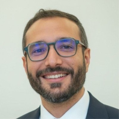 Portrait of Amine Riahi, smiling, wearing glasses and a dark suit